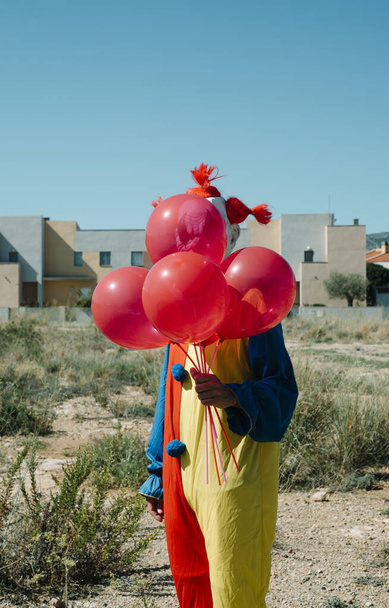 closeup of a creepy clown, wearing a colorful yellow, red and blue costume, is holding a bunch of red balloons in his hand, standing in a vacant lot - Photo, Image