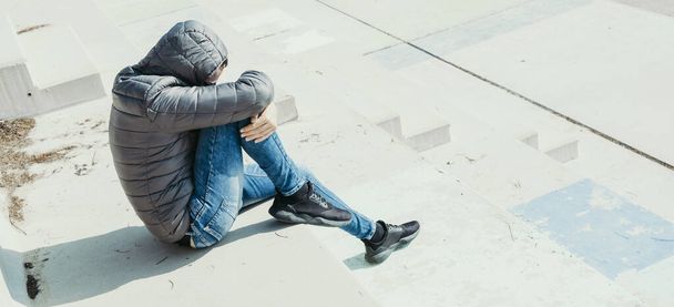 un giovane caucasico, indossando una giacca grigia con cappuccio e jeans, è rannicchiato seduto su una scala esterna in cemento, in un formato panoramico da utilizzare come banner web o intestazione - Foto, immagini