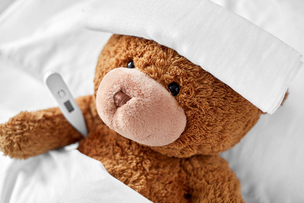 ill teddy bear with bandage and thermometer in bed - Photo, Image