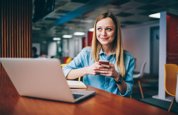 Happy female in casual outfit looking away typing message on smartphone while sitting at wooden table and working in modern workplace - Foto, Imagem