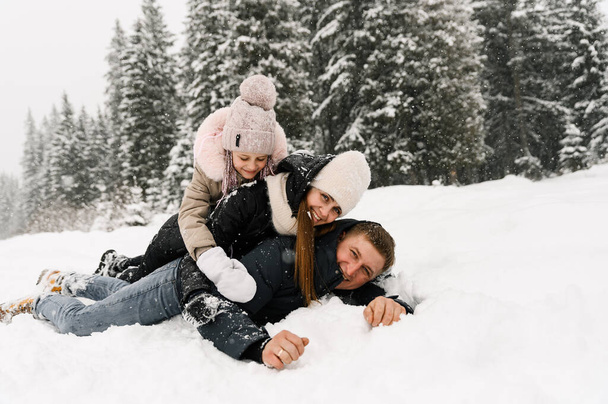 Happy family have fun in winter forest. Mother, father and dauther lie on the ground and playing with snow. Family concept. Enjoying spending time together - Фото, зображення