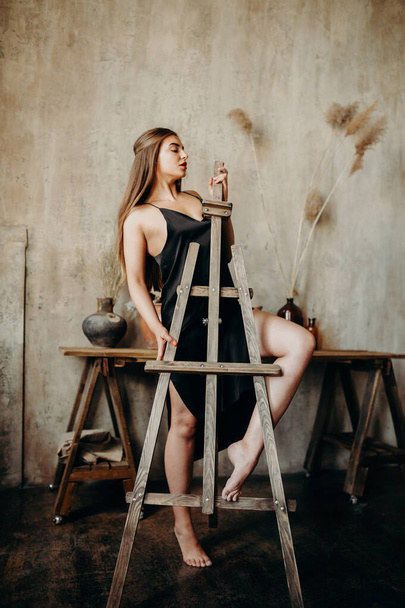 A tall brunette girl in a black dress near a painter's mulbert in an art studio. - Foto, afbeelding