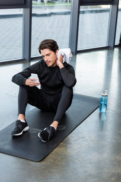 Мужчина пользуется смартфоном и протирает шею инструментом на фитнес-коврике в спортзале  - Фото, изображение