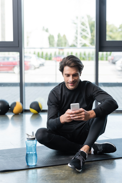 Esportista alegre usando celular perto de garrafa de esportes no tapete de fitness no ginásio  - Foto, Imagem
