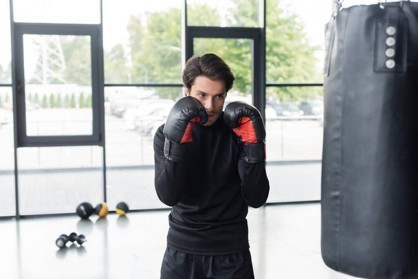 Deportista en guantes de boxeo de pie cerca del saco de boxeo mientras entrena en el gimnasio  - Foto, Imagen