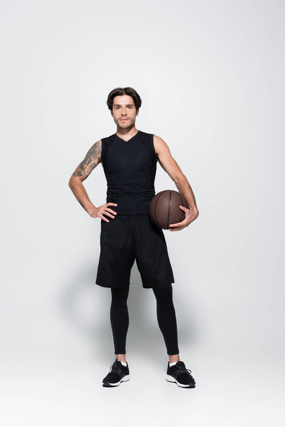 Full length of tattooed player with basketball ball looking at camera on grey background - Foto, afbeelding