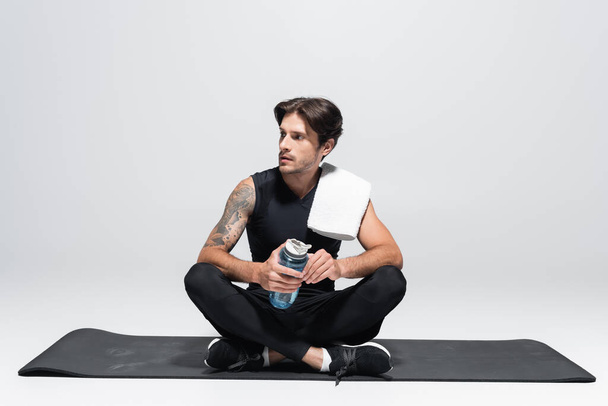 Brunette sportsman with towel and sports bottle sitting on fitness mat on grey background - Photo, Image
