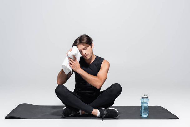 Sportman veegt gezicht met handdoek in de buurt van sportfles op fitness mat op grijze achtergrond - Foto, afbeelding
