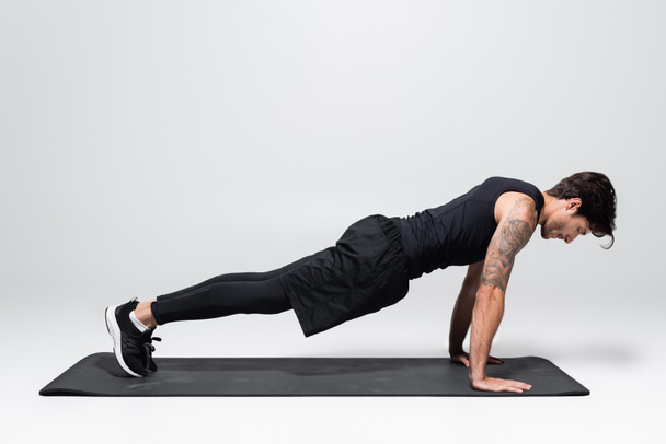 Side view of sportsman in back sportswear doing plank on fitness mat on grey background - Photo, Image