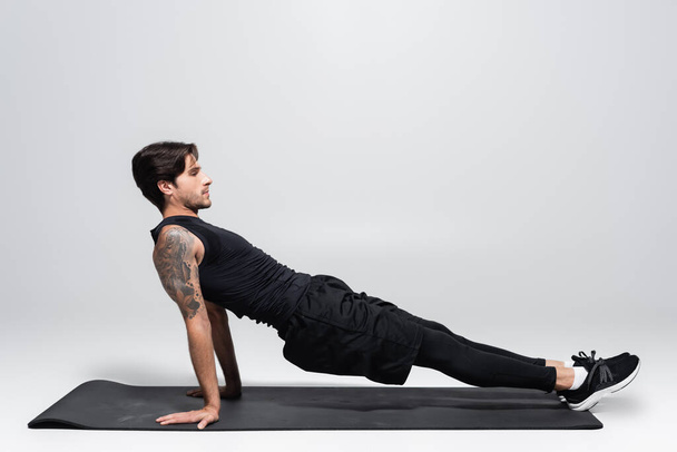 Vue latérale de l'homme faisant planche sur tapis de fitness sur fond gris - Photo, image