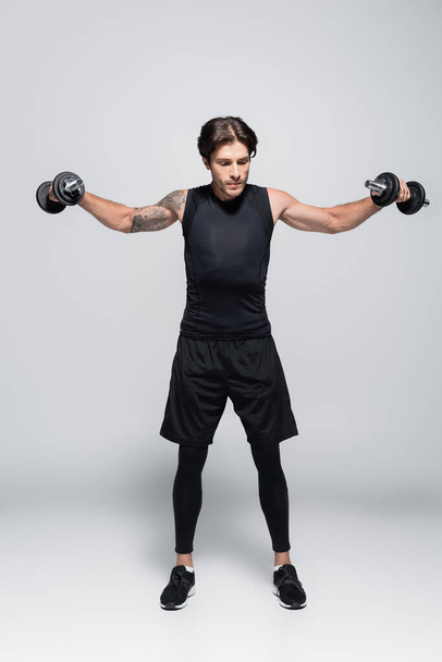 Full length of tattooed sportsman training with dumbbells on grey background  - Photo, Image