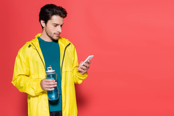 Sportsman in possesso di bottiglia sportiva e utilizzando smartphone su sfondo rosso  - Foto, immagini
