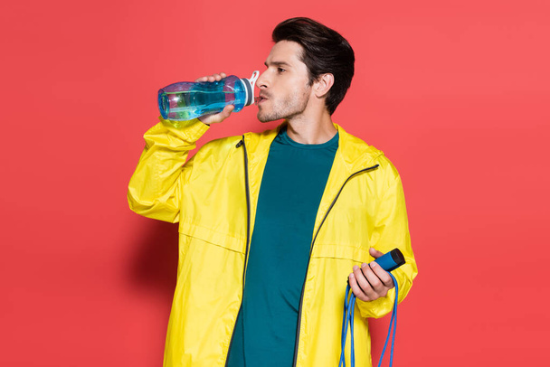 Brunette sportsman holding skipping rope and drinking water on red background  - Foto, immagini