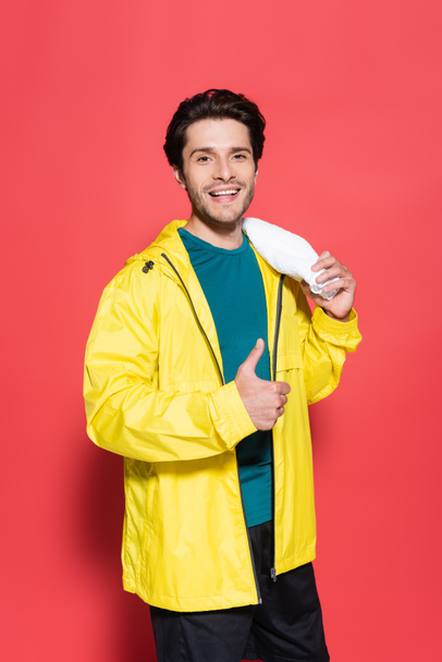 Smiling sportsman in yellow sports jacket showing like and holding towel on red background  - Foto, immagini