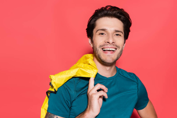 Smiling sportsman holding yellow sports jacket and looking at camera on red background  - Fotografie, Obrázek