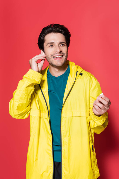 Smiling sportsman in sports jacket holding wireless earphones on red background  - Photo, Image