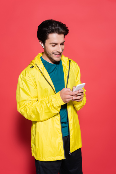 Happy sportsman in earphone using cellphone on red background  - Фото, изображение