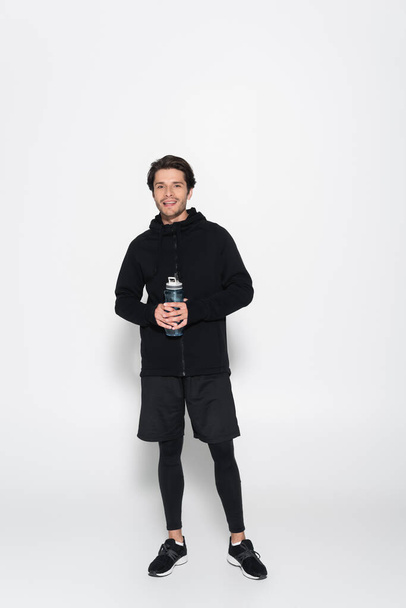 Full length of smiling sportsman holding sports bottle and looking at camera on grey background  - Foto, immagini
