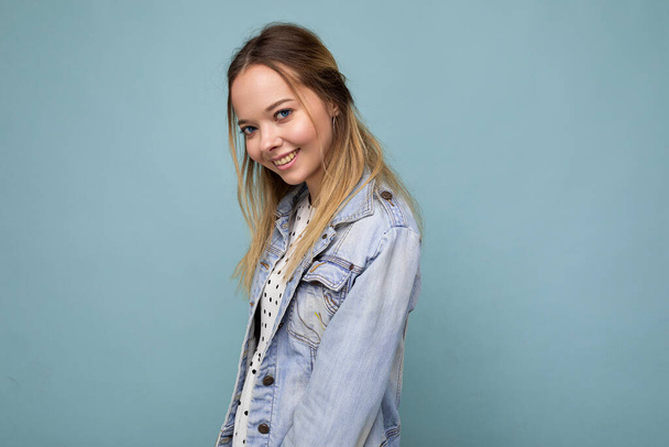 Young beautiful woman. Trendy woman in casual summer dress and stylish denim jacket. Positive female shows facial emotions. Funny model isolated on blue background with free space - 写真・画像