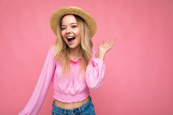 Portrait of young attractive smiling hipster blonde woman in stylish pink crop top and straw hat. Sexy carefree female person posing isolated near pink wall in studio. Positive model with natural - Photo, image