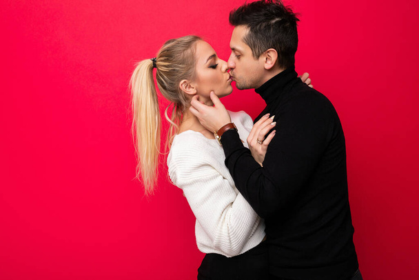 Foto van een mooie jonge blonde vrouw witte gebreide trui en een volwassen brunet man met stoppels in een zwarte trui die geïsoleerd kussen op een roze achtergrond met vrije ruimte voor tekst. Liefde - Foto, afbeelding