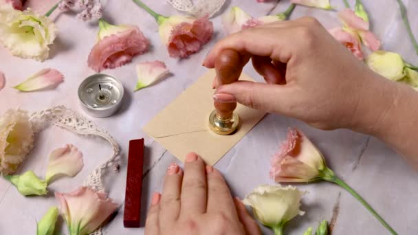 Ręce uszczelniające stopiony wosk na kopercie w pobliżu różowych kwiatów nad marmurowym stołem  - Materiał filmowy, wideo