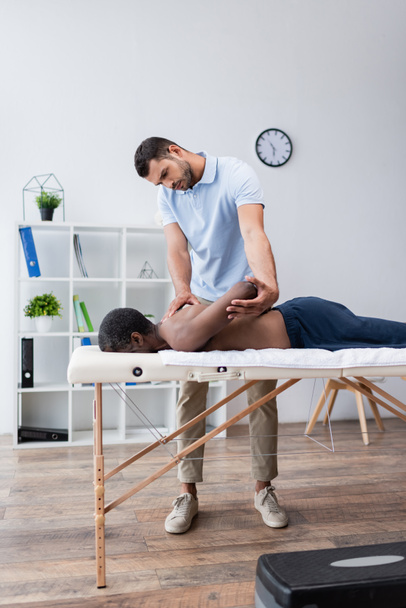 young rehabilitation specialist stretching arm of african american man on massage table - Foto, Imagen
