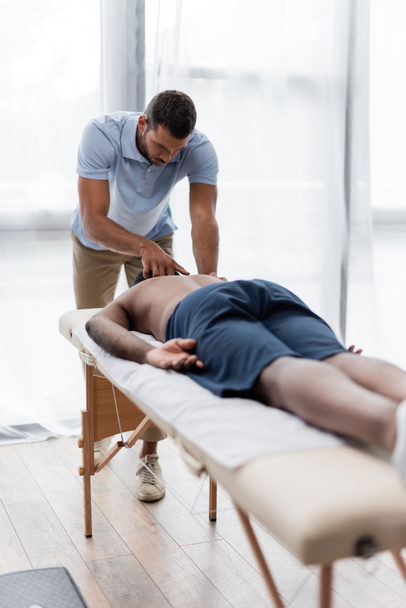 młody fizjoterapeuta robi masaż szyi afrykańskiemu mężczyźnie na niewyraźnym pierwszym planie - Zdjęcie, obraz