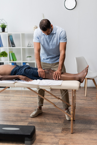 chiropractor massaging knee of african american man in rehabilitation center - Фото, зображення