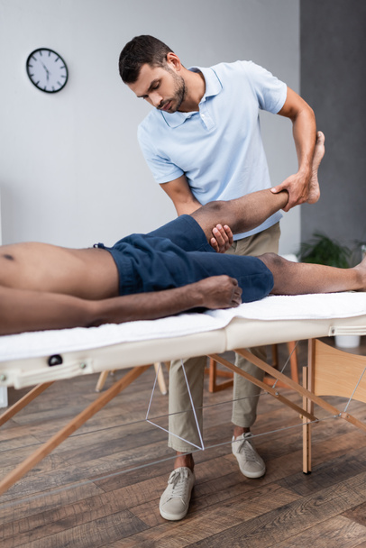 chiropractor massaging leg of african american man in rehabilitation clinic - Fotó, kép