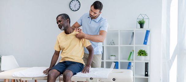 middle aged african american man sitting on massage table while rehabilitologist examining his shoulder, banner - Valokuva, kuva