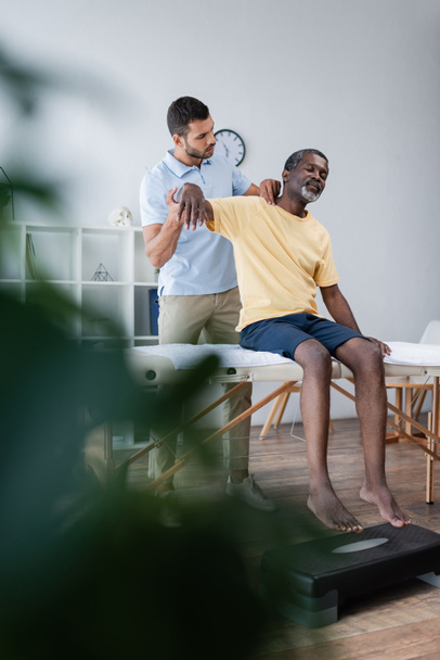 jong fysiotherapeut onderzoeken middelbare leeftijd Afrikaans amerikaanse man op wazig voorgrond - Foto, afbeelding