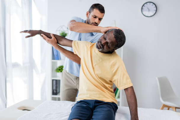 リハビリテーション学者がアフリカ系アメリカ人男性の腕と首をクリニックに伸ばし - 写真・画像