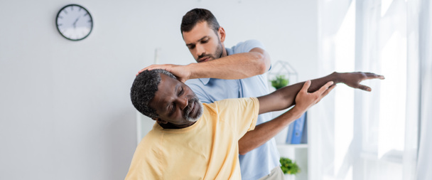 physiotherapist stretching arm of african american man while examining him in rehab center, banner - Foto, Imagen