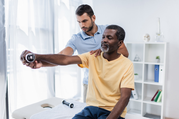 mature african american man working out with dumbbell with help of young rehabilitologist - Foto, Bild