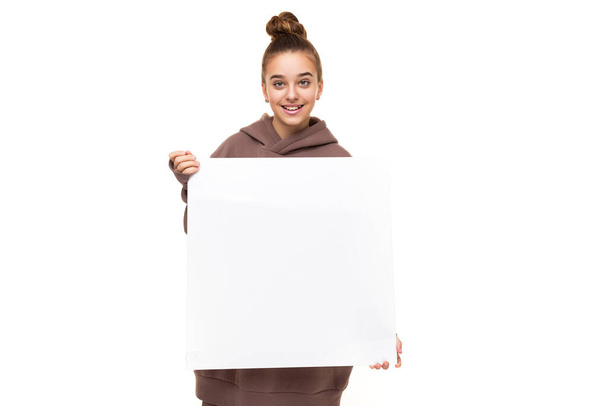 Photo of a beautiful positive smiling cute girl with dark hair and a bun in a brown hoodie with hood holding a large white square sheet with blank space isolated on white background with free space - Photo, image