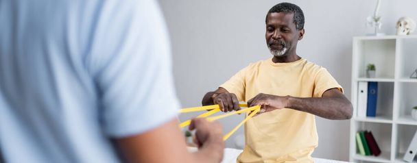 africký Američan cvičí s elastikou spolu s rozmazaným rehabilitologem, bannerem - Fotografie, Obrázek