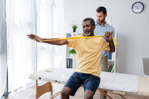 middle aged african american man sitting on massage table and training with elastics near rehabilitologist - Foto, immagini