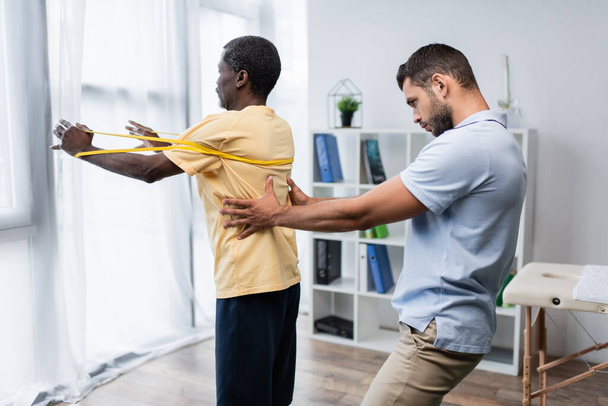 jonge trainer ondersteunen volwassen Afrikaans amerikaanse man uit te werken met elastiek - Foto, afbeelding
