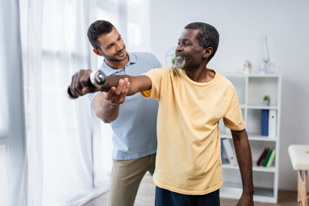 young trainer assisting happy african american man training with dumbbell in rehabilitation center - Fotoğraf, Görsel