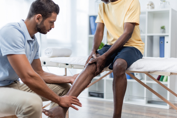 jong rehabilitoloog aanraken been van Afrikaans amerikaanse man zitten op massagetafel - Foto, afbeelding