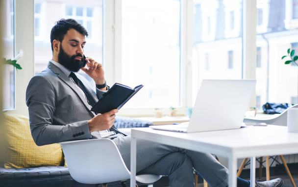 Side view of confident entrepreneur in formal suit sitting at table with laptop while having conversation on smartphone and looking at planner - Φωτογραφία, εικόνα