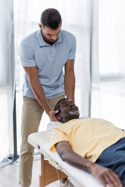 mladý fyzioterapeut léčení zralý afroameričan muž na masážním stole - Fotografie, Obrázek