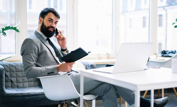 Side view of businessman in formal clothes looking at camera sitting at table with laptop and notebook while having phone call - Φωτογραφία, εικόνα