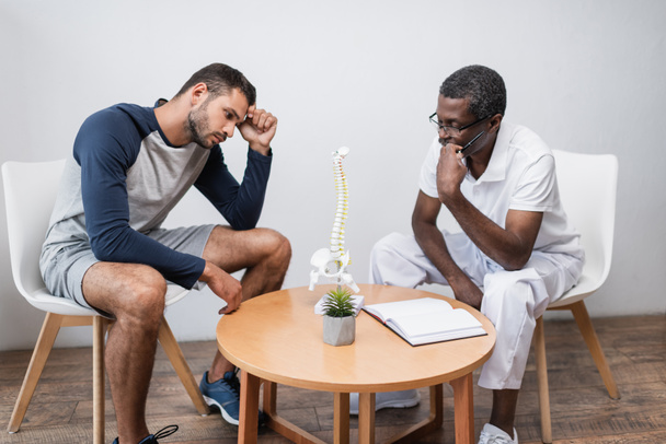 young man and mature african american doctor looking at spine model near blank notebook - Foto, Imagem