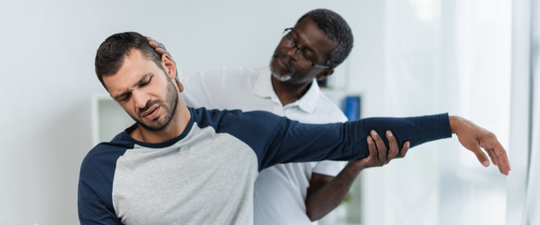 young man frowning from pain while african american rehabilitologist raising his arm, banner - Foto, imagen