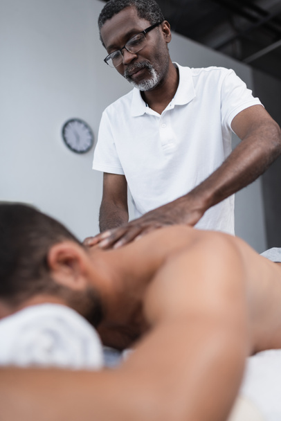vista de bajo ángulo del masajista afroamericano haciendo masaje de espalda durante la rehabilitación del hombre borroso - Foto, imagen