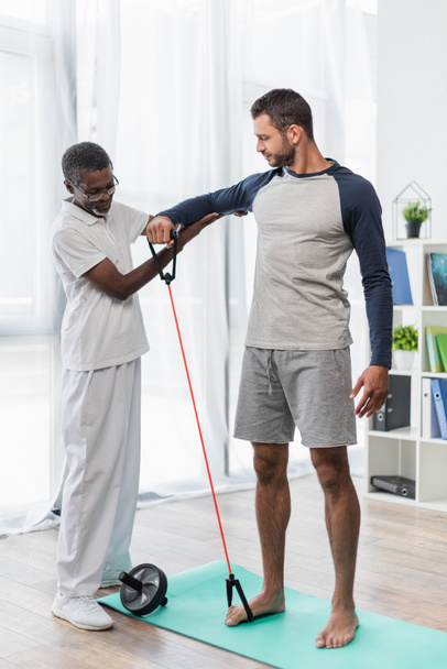 mature african american physical therapist assisting barefoot man working out with resistance band - Foto, afbeelding