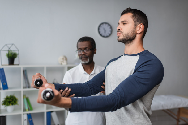 young man exercising with dumbbells near blurred african american rehabilitologist - 写真・画像