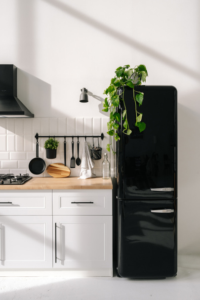 Black shiny refrigerator in scandinavian style kitchen. White cuisine furniture with hanging cutlery. Modern apartment after repair. Stylish household appliance - Zdjęcie, obraz